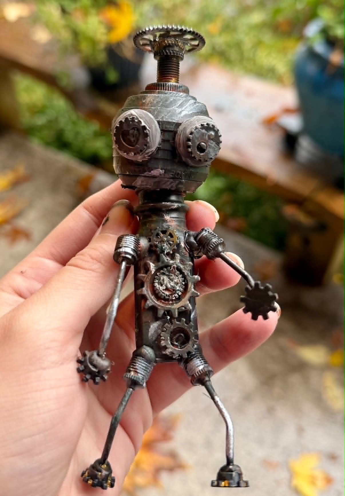 A handmade found-object robot held in a hand outdoors, built from gears and small metal pieces.
